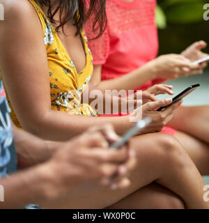 Unposed closeup lifestyle colpo di tre multi-etnico popolo millenario digitando su smartphone ina giardino tropicale con piante verdi Foto Stock