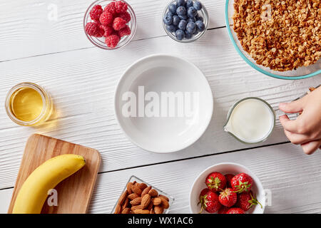 Bacche mature, granola, noci, miele e una piastra vuota su un bianco tavolo di legno. Una donna di mano trattiene un bicchiere di latte. Integratore foo Foto Stock