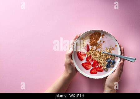 Donna di mani tenendo una tazza di yogurt biologici smoothie con fragole, banana, mirtillo, fiocchi di avena e biscotti rosa su ba Foto Stock