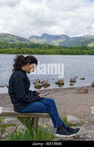 Una giovane donna ignorando il Lake District scenario di concentrarsi sul suo telefono cellulare, Elterwater, REGNO UNITO Foto Stock