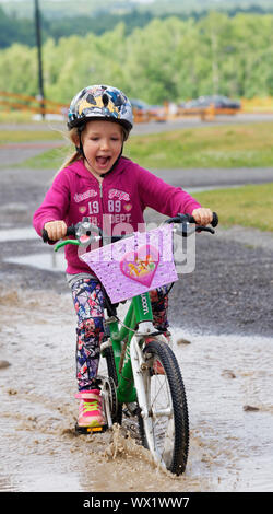 Un sorridente bambina (5 anni) in sella la sua bici attraverso una pozzanghera Foto Stock