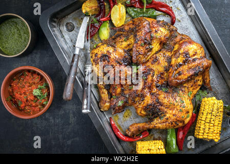 Spatchcocked barbecue pollo al mattone con peperoncino Salsa chimichurri e peperoncino relish come vista dall'alto su un vecchio foglio di metallo Foto Stock