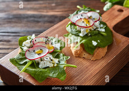 Ciabatta brinda con soffici fiocchi di formaggio, fette di ravanello, spinaci freschi, semi di lino su sfondo di legno. Foto Stock