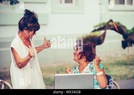 Due entusiasti le donne anziane che celebra il successo durante la lettura di buone notizie online con un computer portatile in outdoor cafe. Foto Stock