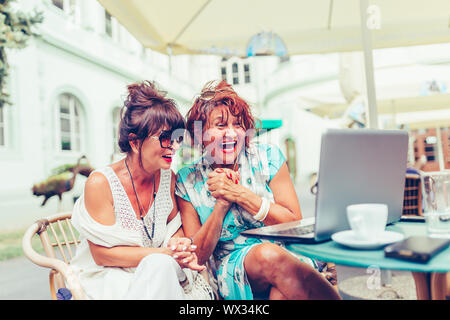 Due entusiasti le donne anziane la lettura di buone notizie on line con un laptop in outdoor cafe Foto Stock