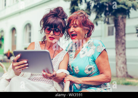 Due entusiasti le donne anziane in seduta outdoor cafe ridendo e guardando i social media e divertenti video su un tablet. Foto Stock