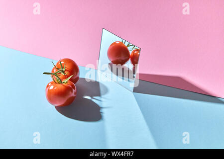 Due pomodori con steli verdi sono riflessa in uno specchio su un Blu-rosa sfondo doppia con copia spazio. Ortaggi sani Foto Stock