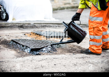 L uomo si riversa di catrame da un annaffiatoio intorno a un chiusino su una strada spianata e riparato nel Regno Unito Foto Stock