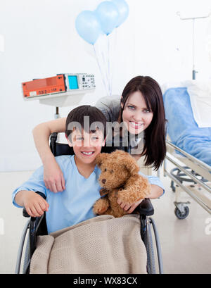 Sorridente piccolo ragazzo seduto su una sedia a rotelle e sua madre in ospedale Foto Stock