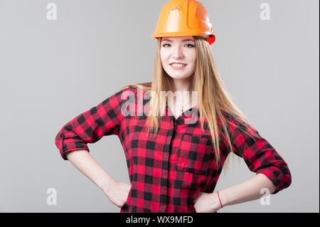 Ritratto di una giovane donna attraente con capelli biondi in arancione casco su uno sfondo grigio neutro. Foto Stock