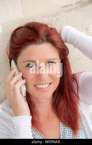 Piuttosto sorridente donna redheaded contro un vecchio muro per effettuare una chiamata con il suo telefono cellulare Foto Stock