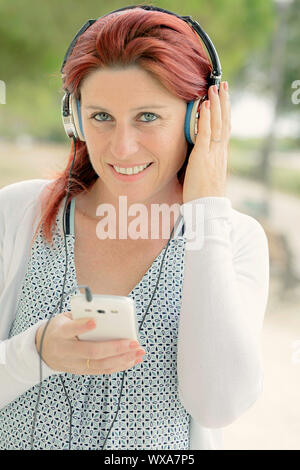 Ritratto di un attraente sorridente redhead donna ascolta musica sul suo telefono con un auricolare con il verde in background Foto Stock