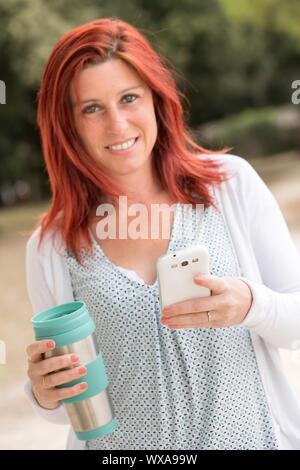Bella sorridente i capelli rossi donna usando un telefono cellulare con una mano e un thermos di caffè nell'altra mano con lo sfondo verde Foto Stock