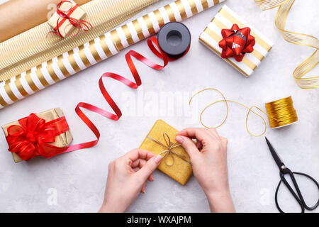 Donna di mani di vincolare il Natale o il regalo di compleanno casella sulla pietra grigia, imbarcazioni carta oro e nastri di colore rosso Foto Stock