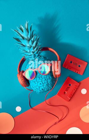 Funny ananas in occhiali da sole e auricolari con un'ombra, piatto stendere sui blu e rosso sfondo della carta Foto Stock