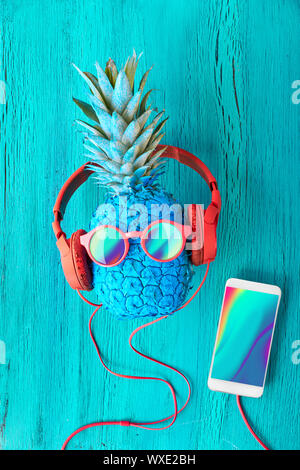 Funny ananas in occhiali da sole e auricolari dipinto di blu. Lay piatto su carta rossa sfondo con sfere di soffici e retrò nastri audio. Foto Stock