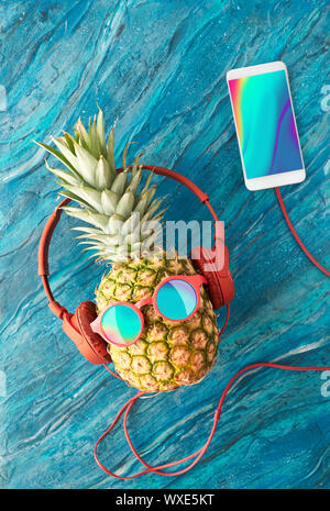 Funny ananas in occhiali da sole e auricolari dipinto di blu. Lay piatto su carta rossa sfondo con sfere di soffici e retrò nastri audio. Foto Stock