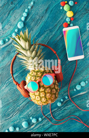 Funny ananas in occhiali da sole e auricolari dipinto di blu. Lay piatto su carta rossa sfondo con sfere di soffici e retrò nastri audio. Foto Stock