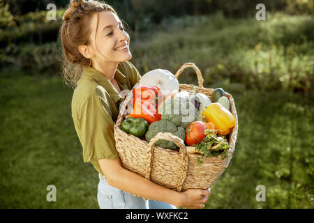 Ritratto di una giovane e bella donna con cesto pieno di appena prelevato ortaggi in piedi nel giardino in una serata di sole Foto Stock