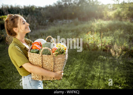 Ritratto di una giovane e bella donna con cesto pieno di appena prelevato ortaggi in piedi nel giardino in una serata di sole Foto Stock