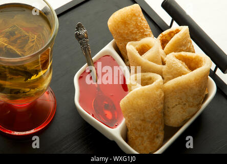 Frittelle con sciroppo e tè al buio su un vassoio. Lussureggiante rosy pancake con sciroppo di luminoso su una piastra bianca e un vecchio cucchiaio accanto a herba Foto Stock