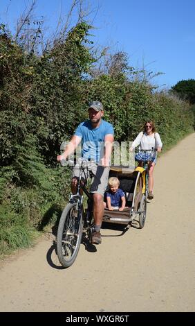 Il padre e la madre tirando little boy in ciclo bambino rimorchio sull'Isola di Sark, Isole del Canale, UK. Foto Stock