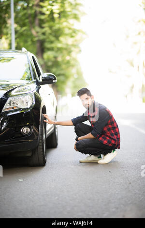 Allegro giovane imprenditore ha problemi con il volante della sua vettura. Egli è kneeing e guardando con serietà Foto Stock