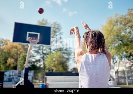 Foto dal retro dell'atleta gettando palla in Basketball hoop contro il cielo blu sul giorno di estate Foto Stock