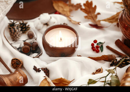 Hygge lifestyle. Candela, bacche e foglie di autunno, erbe e ghiande, dadi e marrone cappello sul tessuto bianco. In autunno l'umore. Ciao autunno, accogliente immagine ispiratrice. Foto Stock