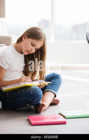 Ragazza di scrittura o di disegno in notebook sedersi sul pavimento. La ragazza non compiti a casa. Foto Stock