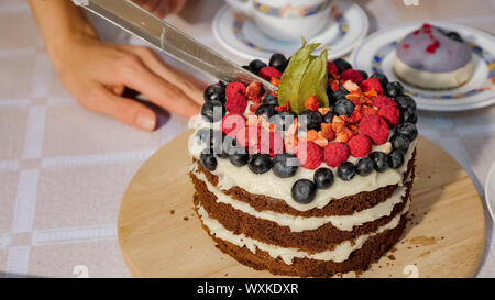 Donna taglia un pezzo di compleanno in casa torta al cioccolato con panna e torte a base di pan di spagna decorata strati di frutti di bosco freschi, primo piano. Foto Stock