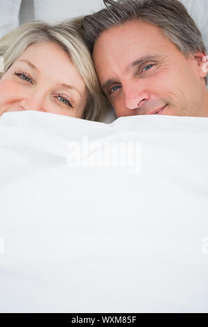 Coppia sorridente in telecamera da sotto le coperte a casa nel letto Foto Stock