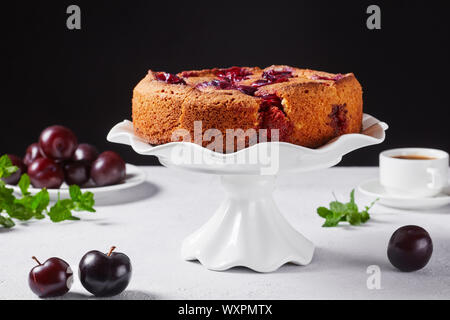 Famosa torta di prugne su una torta bianca stand con prugne fresche su una tavola con fondo nero, spazio libero, close-up Foto Stock