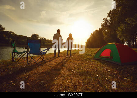 Un escursionista giovane tenendo le mani accanto a una tenda in riva al lago al tramonto. Foto Stock