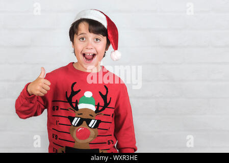 Bambino sorridente indossando Christmas Santa Claus Hat mostra pollice in alto su Sfondo mattone Foto Stock