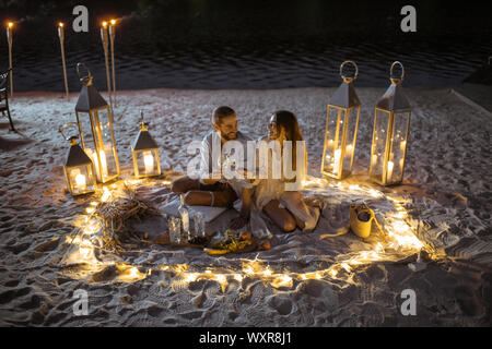 Coppia avente una cena romantica, avvolgente e il tintinnio dei bicchieri da vino presso il splendidamente decorate luogo illuminato con luci diverse la spiaggia di sabbia al tramonto Foto Stock