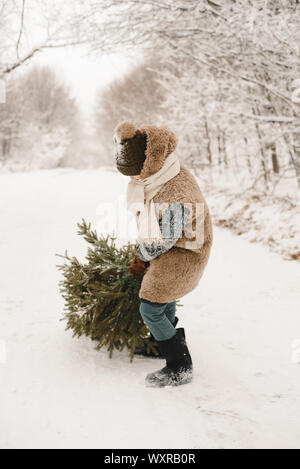 Un piccolo ragazzo porta un albero di Natale in un elfo costume in una foresta innevata. Una nana in una pelliccia è trascinando un albero lungo una strada innevata. Foto Stock