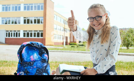 A undici anni da studentessa con un libro si siede sul prato e mostra un dito in alto. Buon umore. Foto Stock