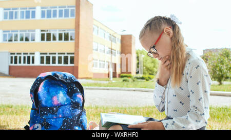 11 enne schoolgirl legge un libro mentre seduto sull'erba sul prato vicino alla scuola. Foto Stock