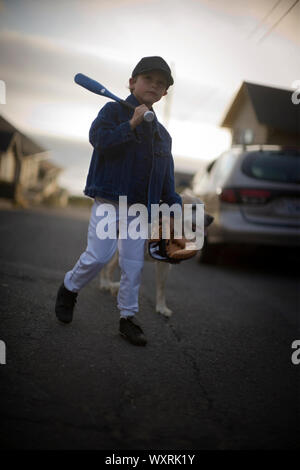 Ritratto di un giovane ragazzo che porta una mazza da baseball e guanto mentre si cammina giù per una strada con il suo cane. Foto Stock