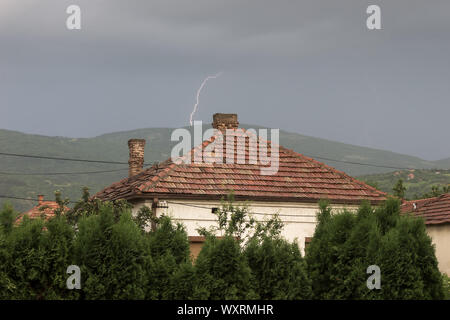 Distant Thunder colpendo la parte superiore della collina dietro il tetto della casa con alberi in primo piano e cavi di energia elettrica durante un temporale Foto Stock