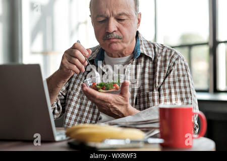 Senior uomo ispanico mangiare insalata fresca al mattino in salotto vicino al laptop Foto Stock