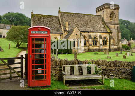 Telefono rosso stand presso medieval San Barnaba chiesa anglicana con il cimitero e il muro di pietra porta umido in condizioni di tempo piovoso in Snowshill Cotswold Inghilterra Foto Stock