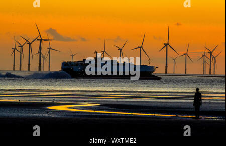 Una nave passa come il sole tramonta dietro Burbo Bank wind farm e 'un altro luogo", un arte di installazione da Anthony Gormley, a Crosby Beach in Merseyside nel nord ovest dell'Inghilterra. Foto Stock