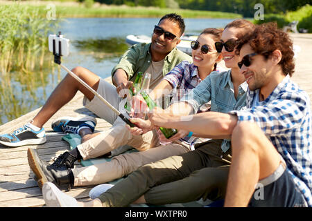 Tempo libero, picnic e concetto tecnologico - amici con bevande di scattare una foto da selfie stick sul lago di pier in estate park Foto Stock