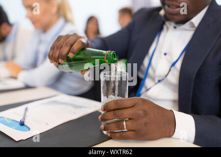 Aziende e persone concetto - close up di African American businessman versando acqua dalla bottiglia di vetro alla conferenza Foto Stock