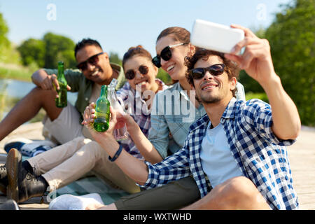Tempo libero, picnic e concetto tecnologico - amici con drink prendendo selfie tramite smartphone sul lago di pier in estate park Foto Stock