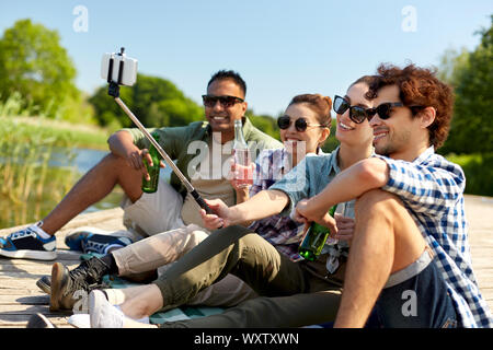Tempo libero, picnic e concetto tecnologico - amici con bevande di scattare una foto da selfie stick sul lago di pier in estate park Foto Stock