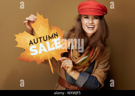 Felice alla moda di 40 anno vecchia donna nel cappuccio rosso, sciarpa, jeans e maglietta cardigan mostra autunno vendita banner contro sfondo beige. Foto Stock