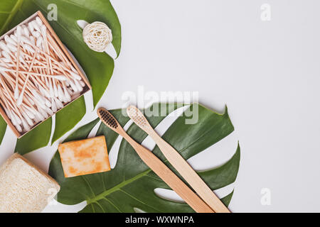 Rifiuti Zero bagno essentials, top voew. Il bambù spazzolini da denti, tamponi di cotone e altri sul tavolo bianco con foglie di monstera Foto Stock